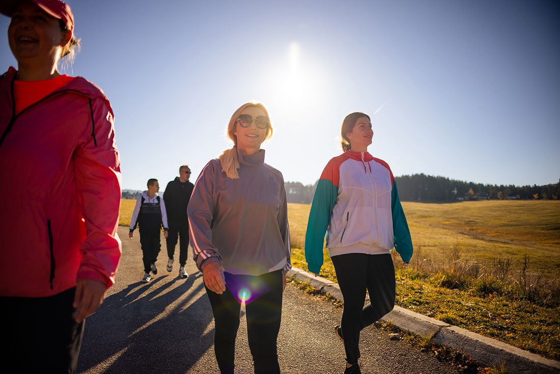 Im Vordergrund gehen drei Personen nebeneinander auf einem asphaltierten Weg, weitere Personen folgen im Hintergrund. Sie befinden sich in einer offenen Feldlandschaft bei tief stehender Sonne und klarem Himmel.