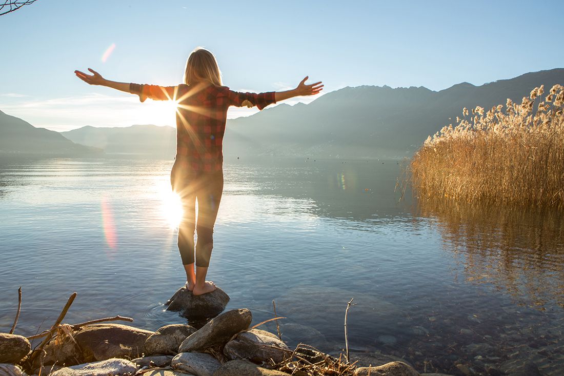Person steht barfuß auf einem Stein am See, mit ausgebreiteten Armen vor Sonne und Bergen.