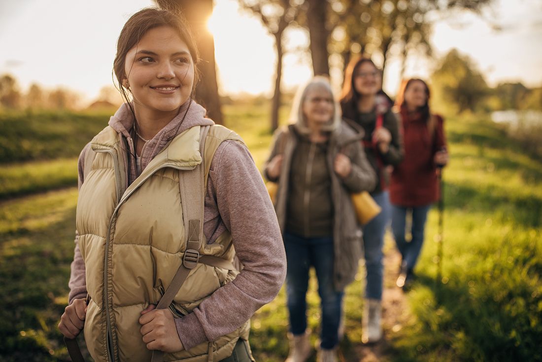 Im Vordergrund geht eine Frau mit Rucksack auf einem schmalen Weg, dahinter folgen drei weitere Frauen. Sie bewegen sich durch eine grüne Landschaft mit Bäumen im warmen Sonnenlicht.