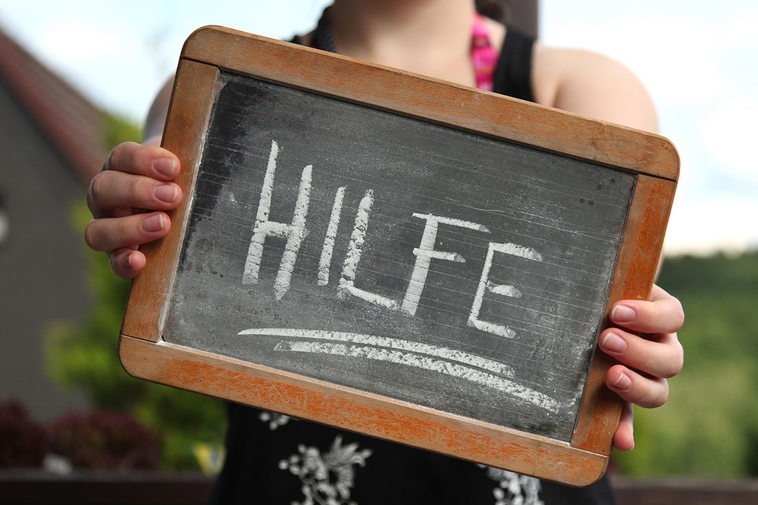 Person hält eine Tafel mit dem Wort „HILFE“ in Kreide vor unscharfem Außenhintergrund.