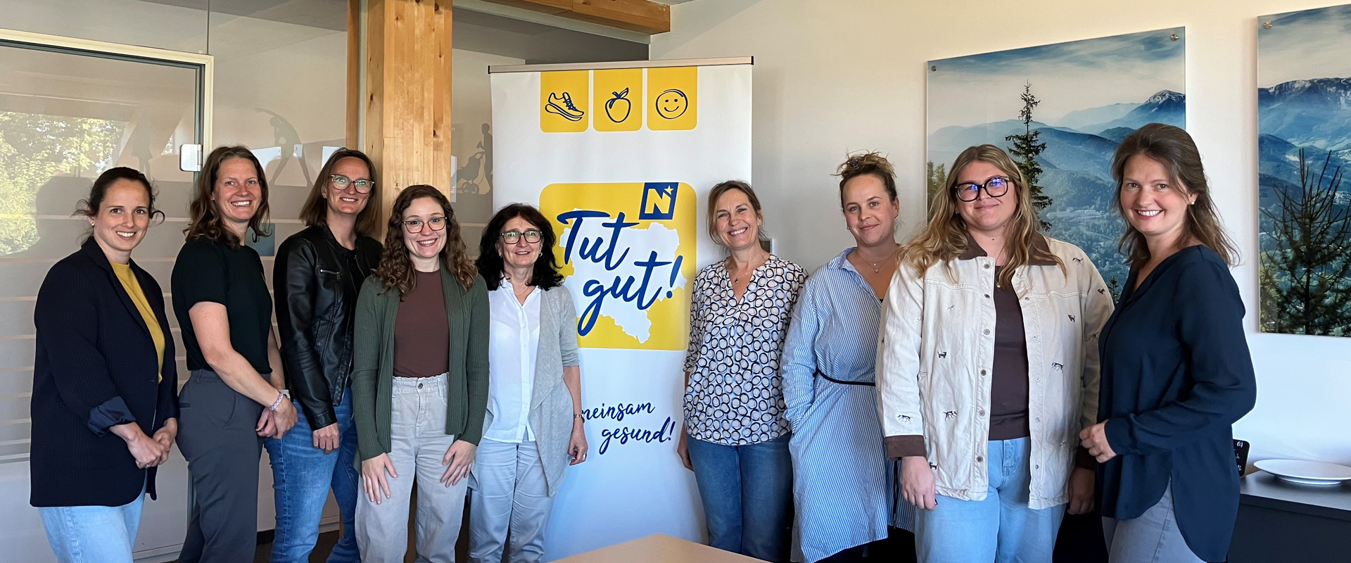Neun Personen stehen für ein Gruppenfoto in einem hellen Besprechungsraum. In der Mitte steht ein Banner mit dem Text „Tut gut!“ und „gemeinsam gesund!“, rechts hängen Landschaftsbilder an der Wand.