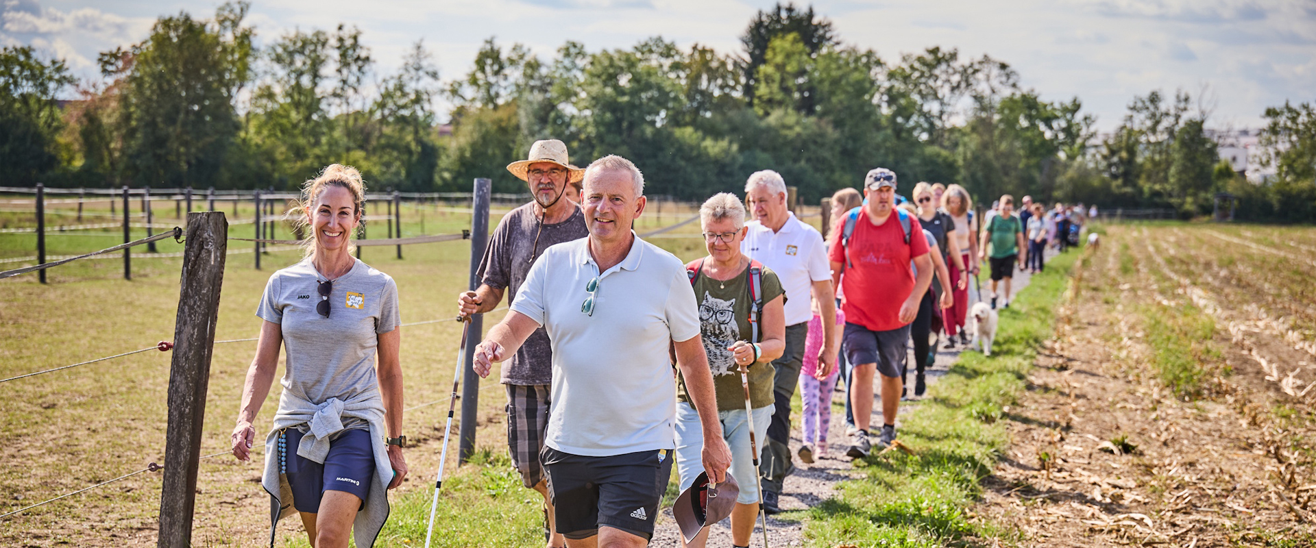 Eine größere Gruppe von Erwachsenen und Kindern wandert auf einem schmalen Weg durch eine ländliche Umgebung. Links verläuft ein Zaun, rechts liegt ein abgeerntetes Feld.