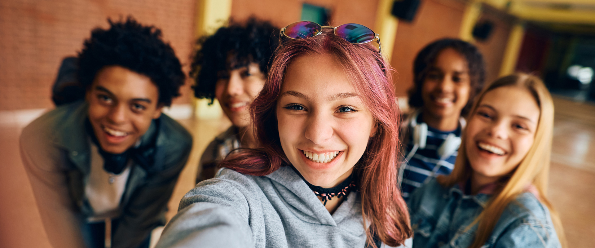 Fünf Jugendliche sitzen nah beieinander und blicken lächelnd in die Kamera. Eine Person im Vordergrund hält den Arm nach vorne, um ein Selfie aufzunehmen.