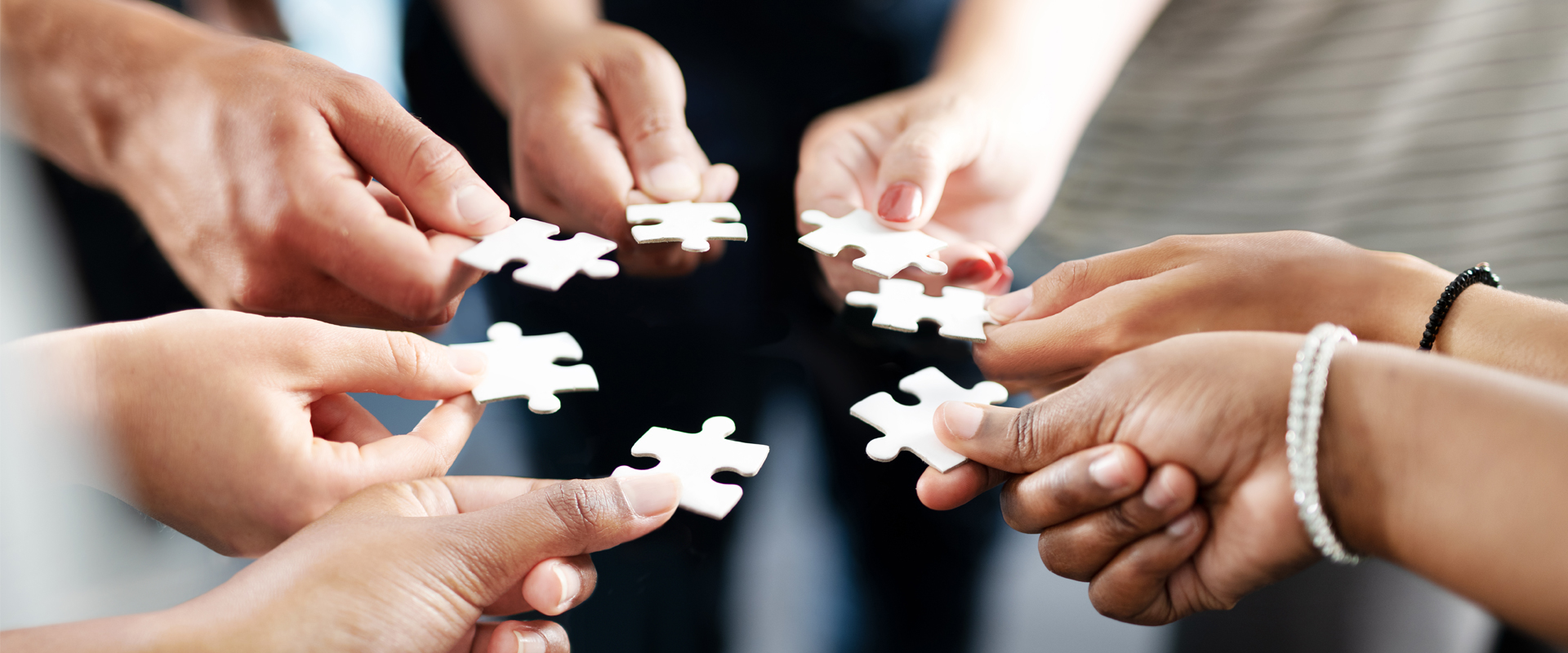 Mehrere Personen halten einzelne weiße Puzzleteile mit den Händen zur Mitte hin. Die Puzzleteile sind noch nicht zusammengesetzt; der Hintergrund ist unscharf.