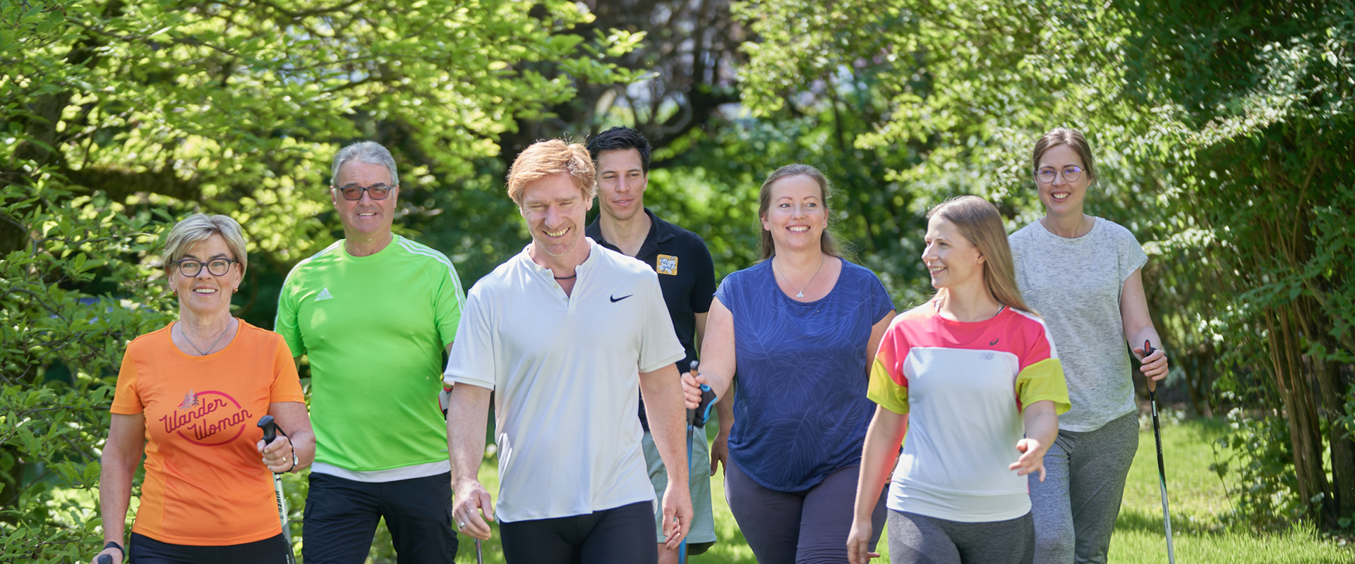 Acht Personen gehen mit Nordic-Walking-Stöcken über eine Wiese im Grünen. Eine gemischte Gruppe aus acht Personen geht mit Nordic-Walking-Stöcken über eine grüne Wiese. Im Hintergrund sind Bäume und dichtes Blattwerk zu sehen.