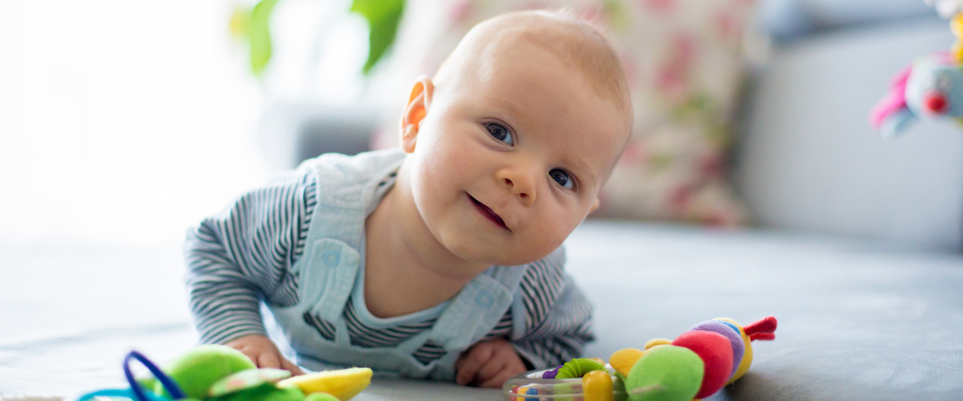 Ein Baby liegt auf dem Bauch auf einer hellen Fläche und blickt in die Kamera. Vor ihm liegen mehrere bunte Spielzeuge, der Hintergrund ist hell und unscharf.