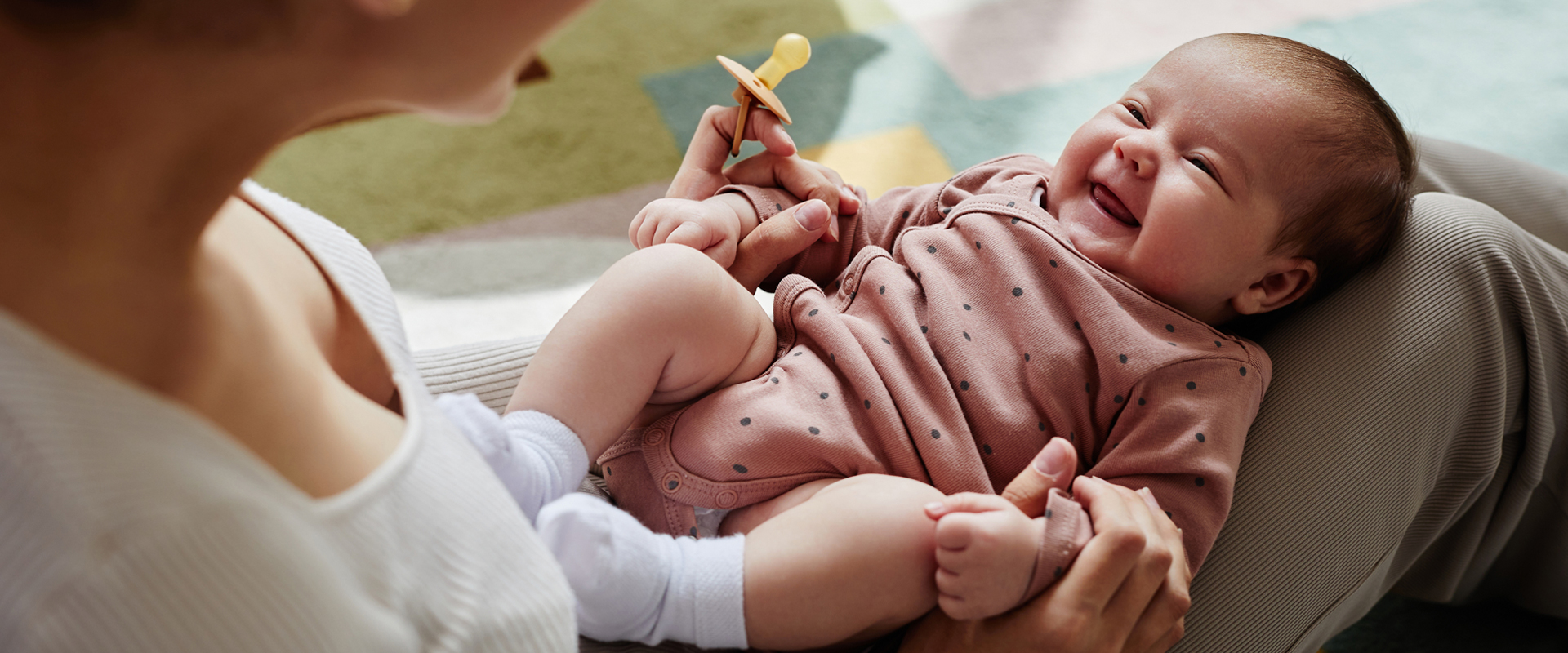 Eine erwachsene Person sitzt mit einem Baby auf dem Schoß und hält es mit beiden Händen. Das Baby liegt auf dem Rücken, lacht und hält einen Schnuller in der Hand.
