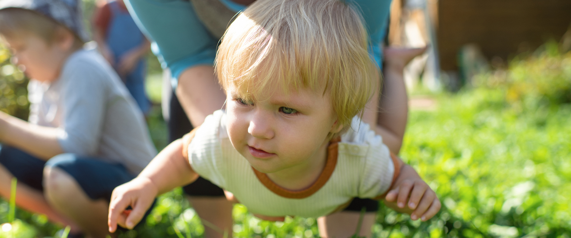 Ein blondes Kleinkind krabbelt auf einer Wiese und wird von einer erwachsenen Person an den Seiten gehalten. Im unscharfen Hintergrund sitzt ein weiteres Kind im Gras.